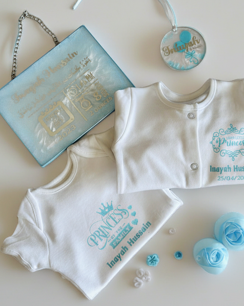 Baby clothing set with 'Prince' text  next to a baby birth stats on a white and blue glitter rectangular shape place with a silver chain for hanging and a ornament with a name on a light background