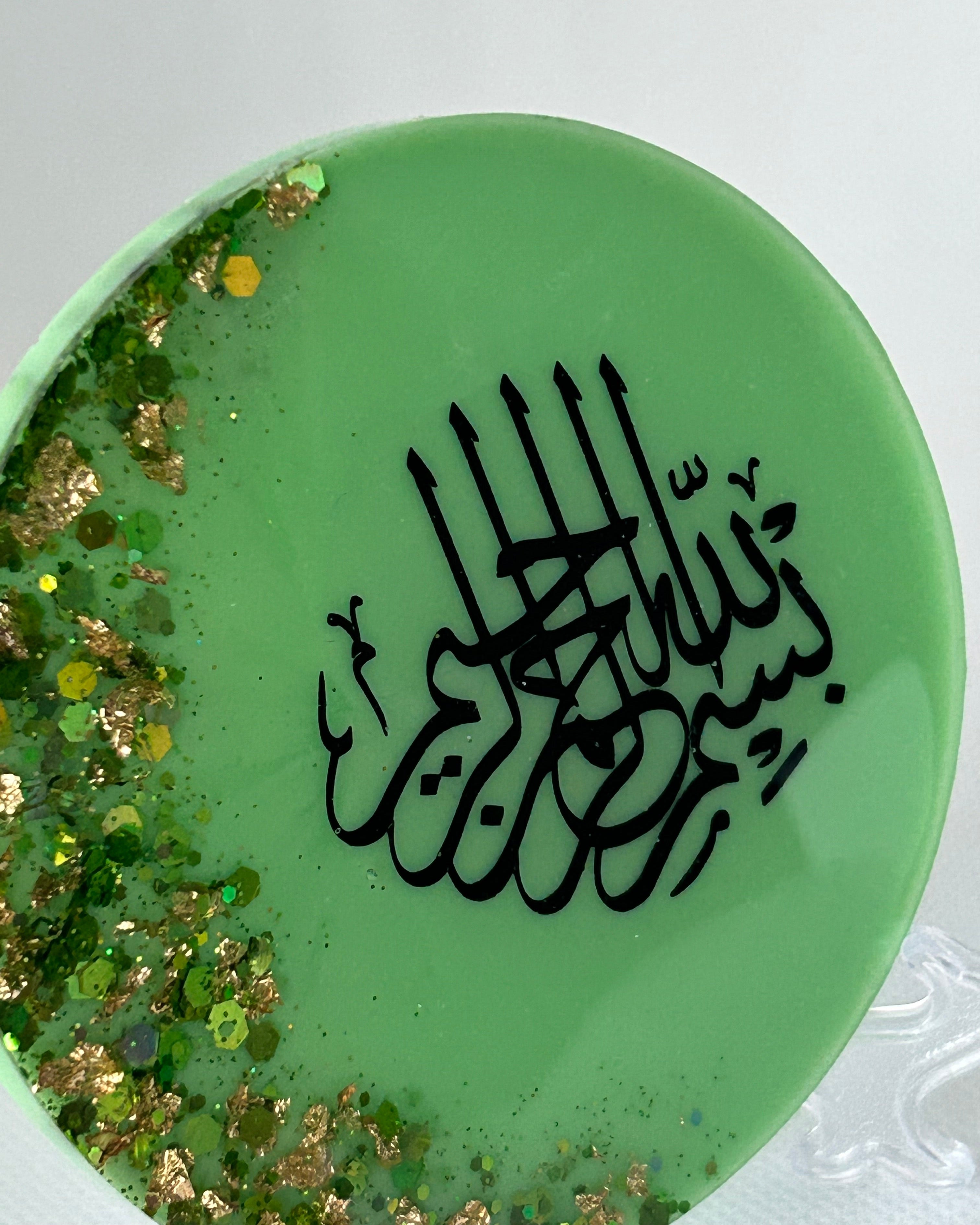 Green circular object with black Arabic calligraphy reading Bismillah Tasmiyah and gold shimmer glitter with gold leaf foiled placed sparsely displayed on a clear plastic display stand on a white background