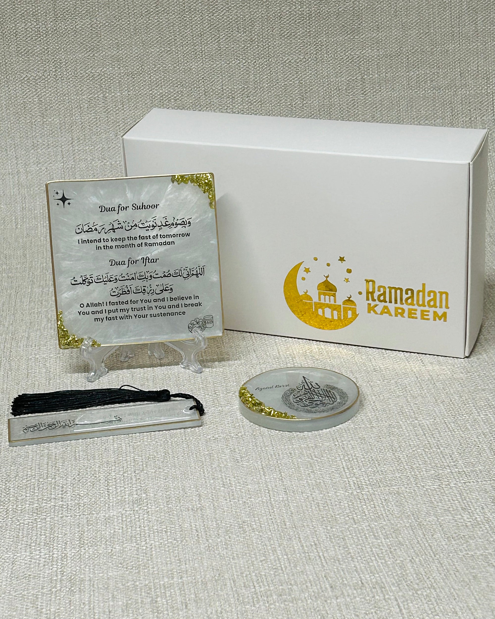Set of items including a white craft box with Ramadan Kareem in gold, a small plaque with Ramdan suhoor and iftar dua, and Ayatul Kursi fridge magnet ans a Quran bookmark with Bismillah in Arabic on a textured surface.