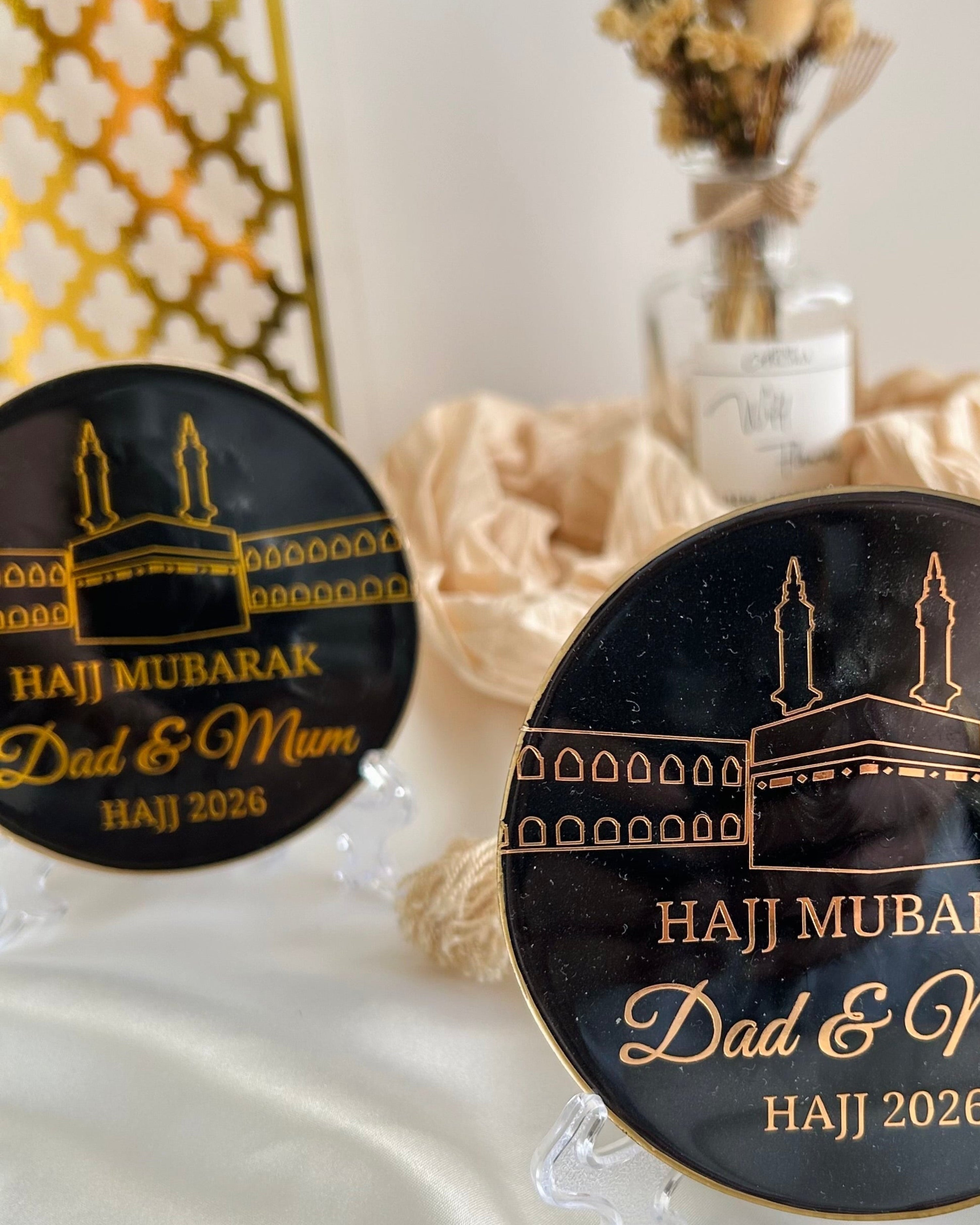 Two black decorative black plaques with 'Haji Mubarak Dad & Mum' text on a stand, featuring a religious symbol 'Kaaba' on a white satin background with decorative items.