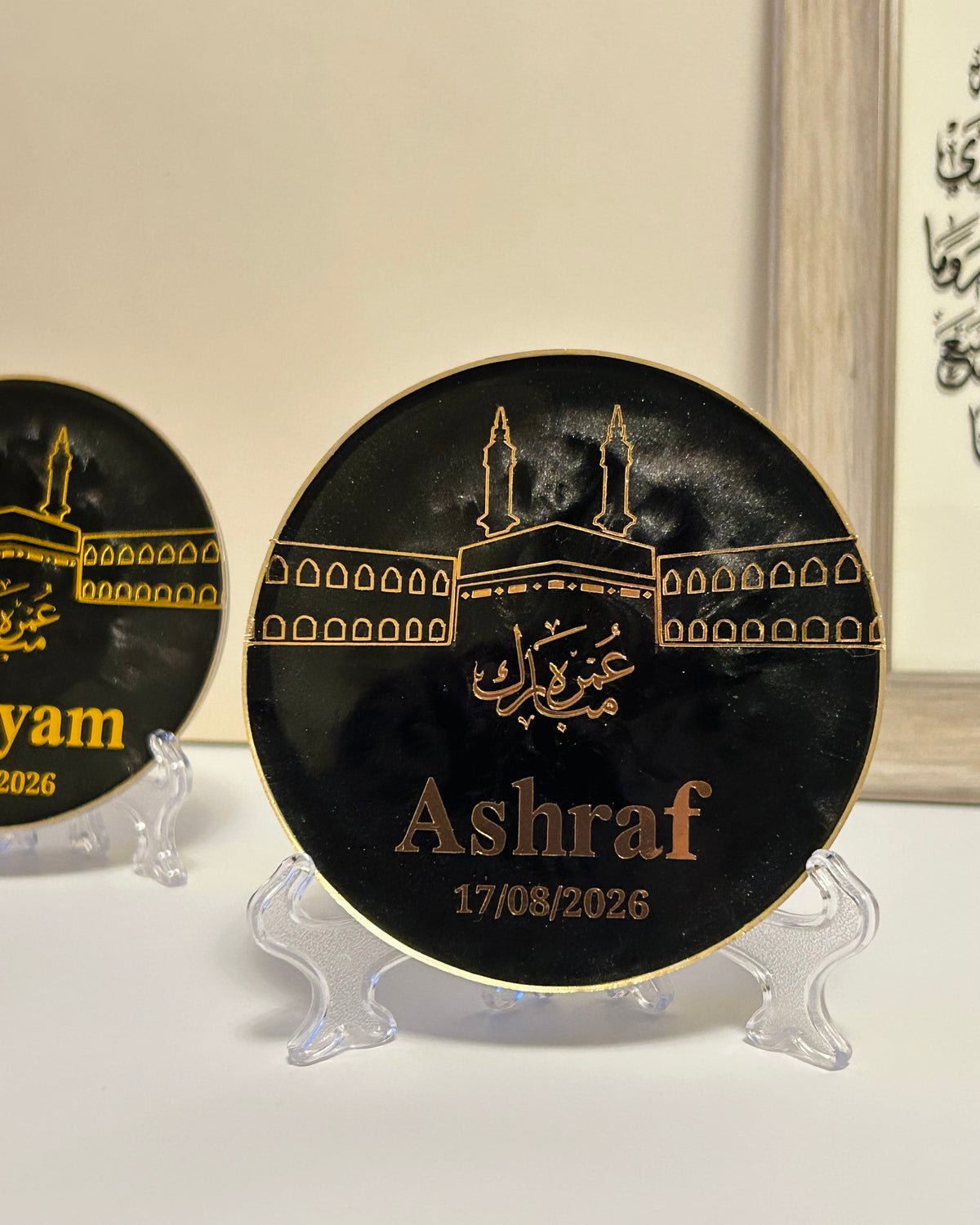 A round black commemorative plaque with gold Arabic calligraphy and a gold Kaaba illustration, displayed on a clear stand. The plaque is personalized with the name 'Ashraf' and the date '17/08/2026'.