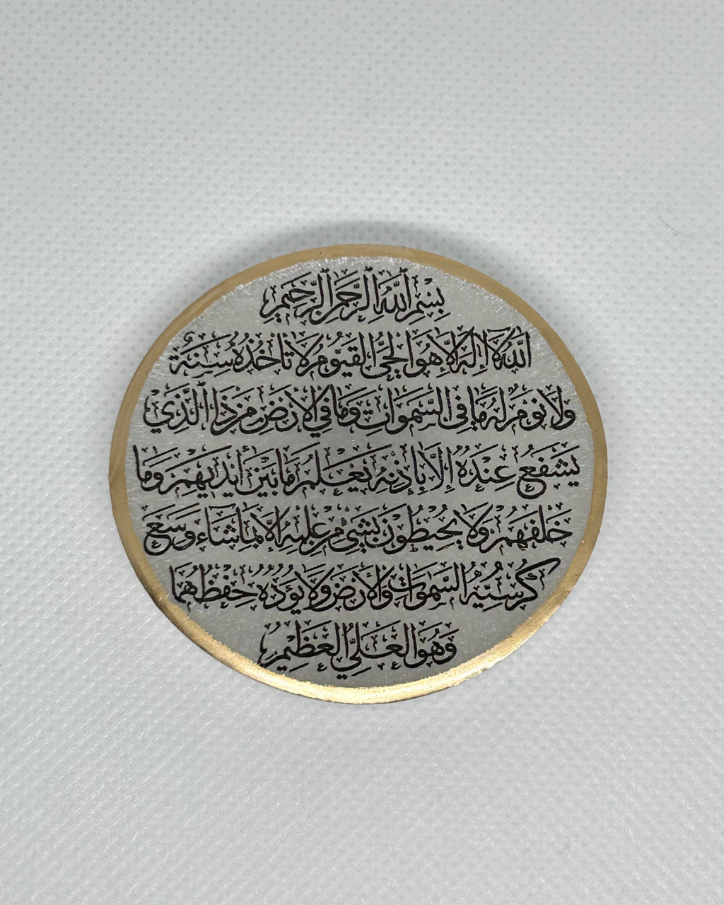 Round white metallic object with Arabic text Ayaytul Kursi in black text and gold decorative edgees on a white background