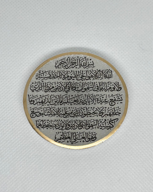 Round white metallic object with Arabic text Ayaytul Kursi in black text and gold decorative edgees on a white background