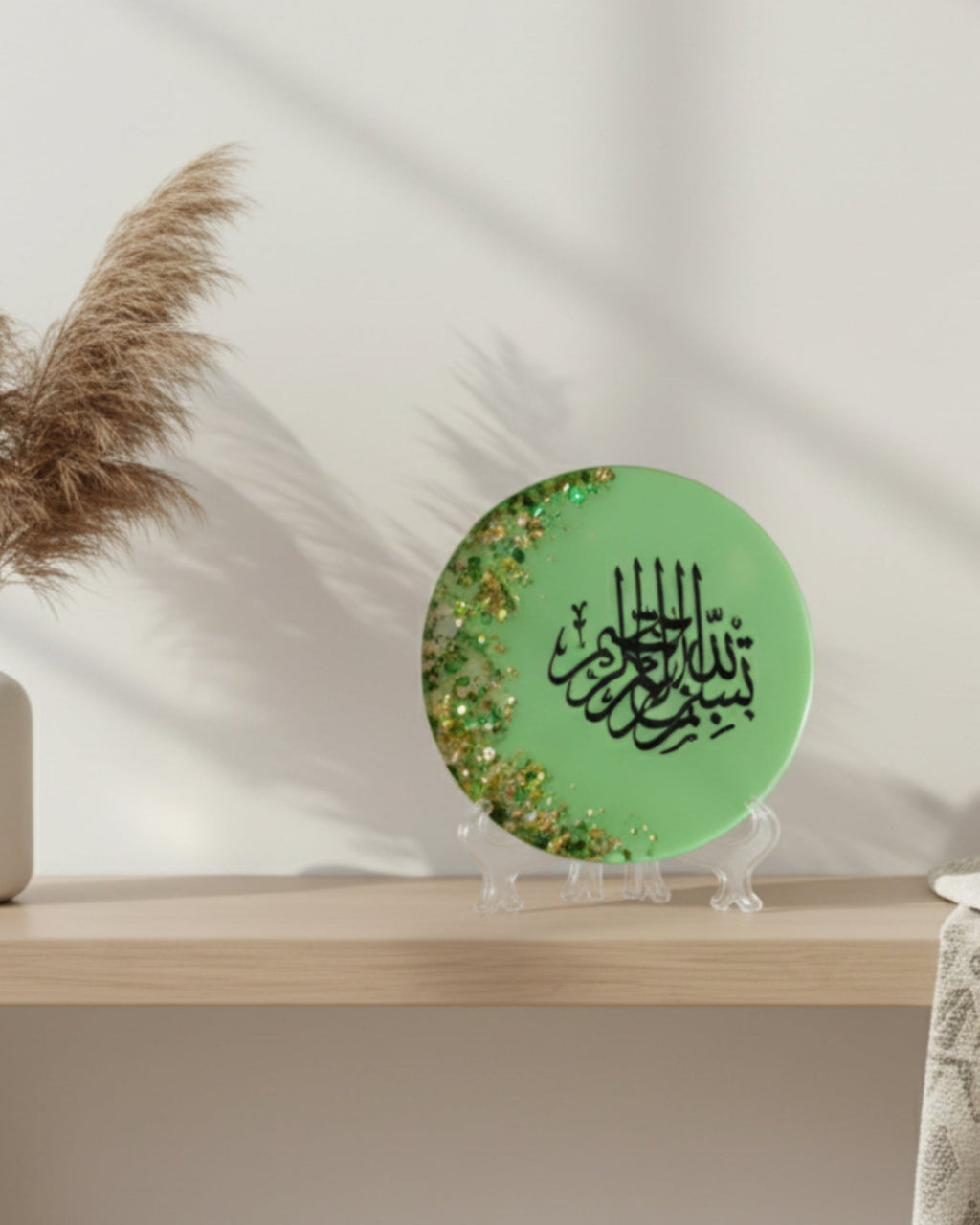 Green circular object with black Arabic calligraphy reading Bismillah Tasmiyah and gold shimmer glitter with gold leaf foiled placed sparsely displayed on a clear plastic display stand on a white background