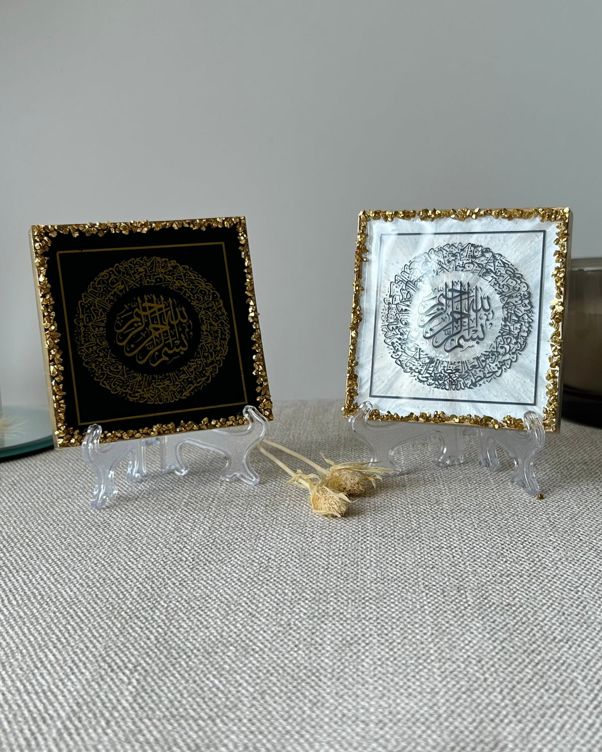 Two square Islamic plaques with Ayatul Kursi in Arabic calligraphy, displayed on clear acrylic stands on a textured beige fabric surface. The left plaque has a black background with gold crushed border and gold calligraphy, the right plaque as a pearlescent white background with gold crushed glass border and black calligraphy. Both plaques are displayed on frosted plastic display stands.