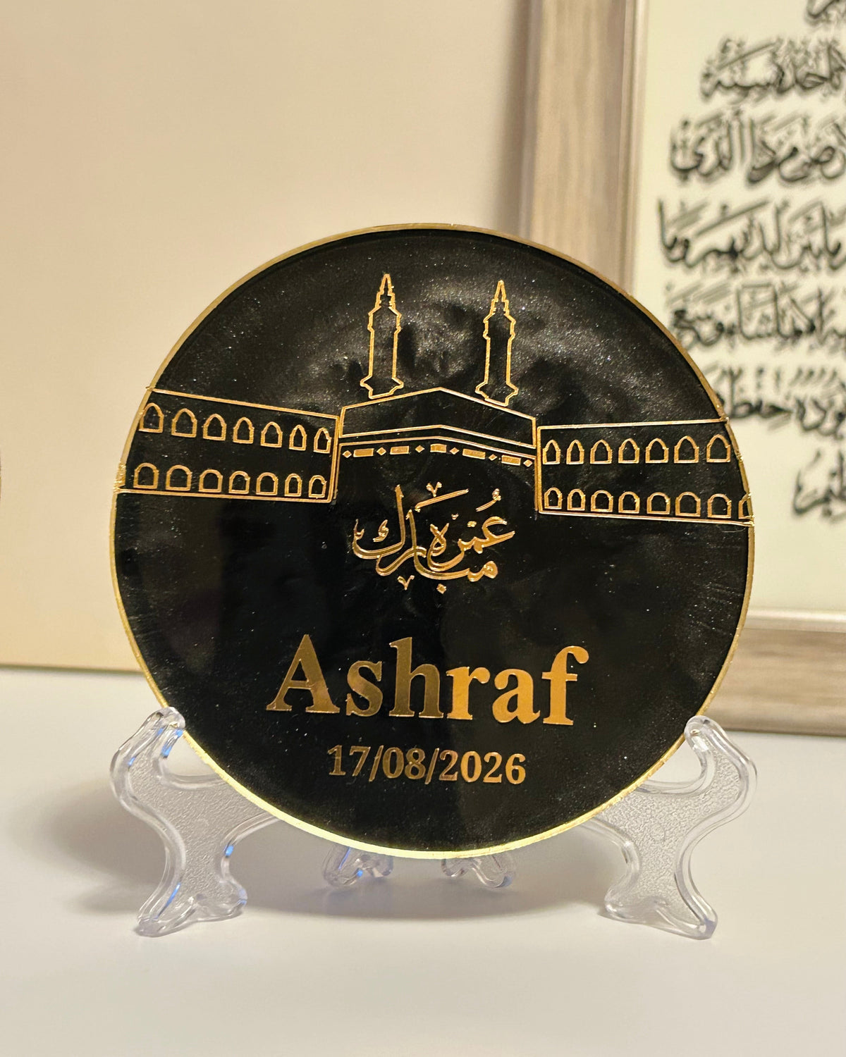 A round black commemorative plaque with gold Islamic artwork and the name 'Ashraf' plus the date '17/08/2026', displayed on a clear stand against a light indoor background.
