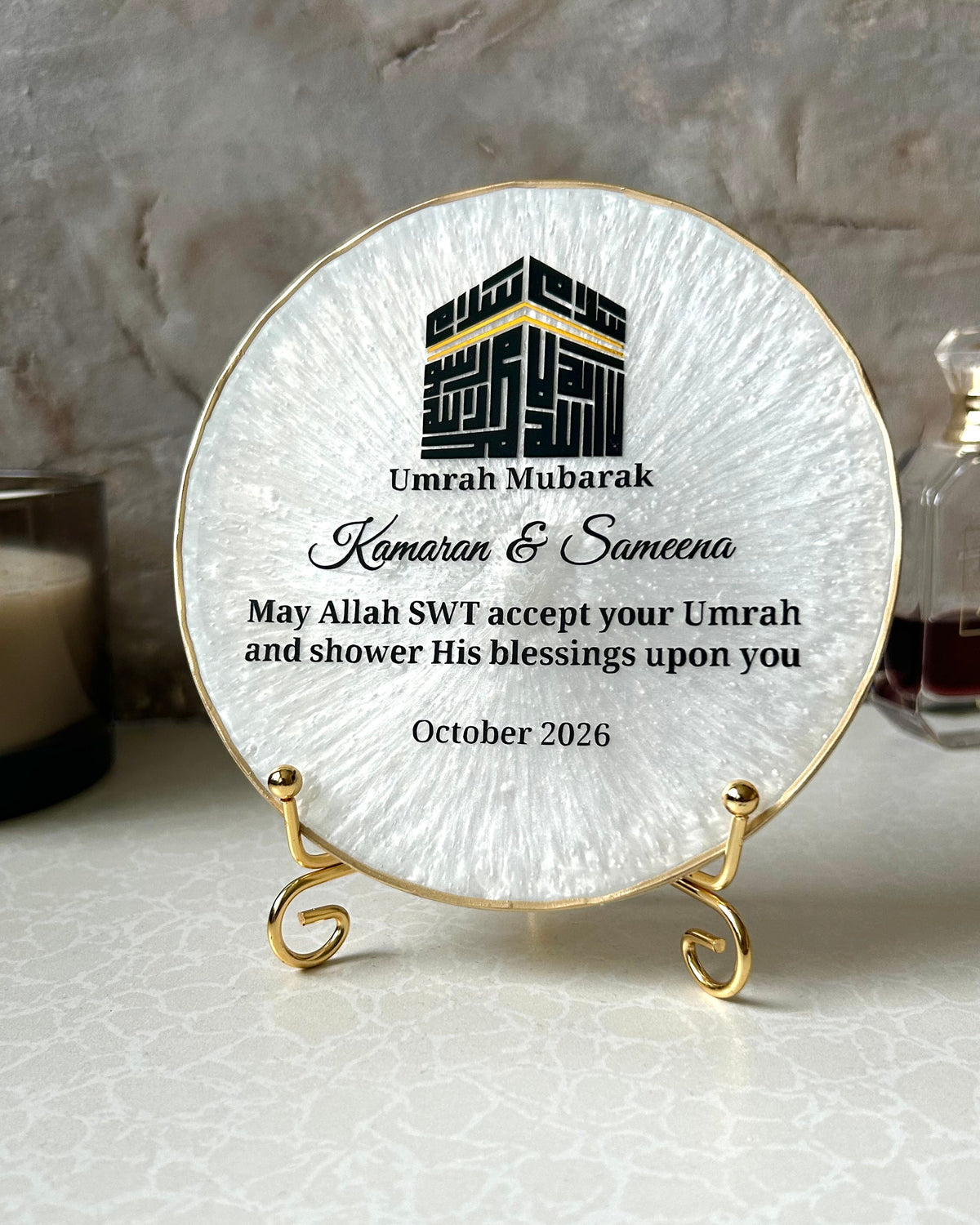 A round pearlescent white resin plaque with a gold rim and gold display stand, with a black Kufic Kaaba illustration reading Basmala and personalised Umrah Mubarak text, displayed on a marble surface.