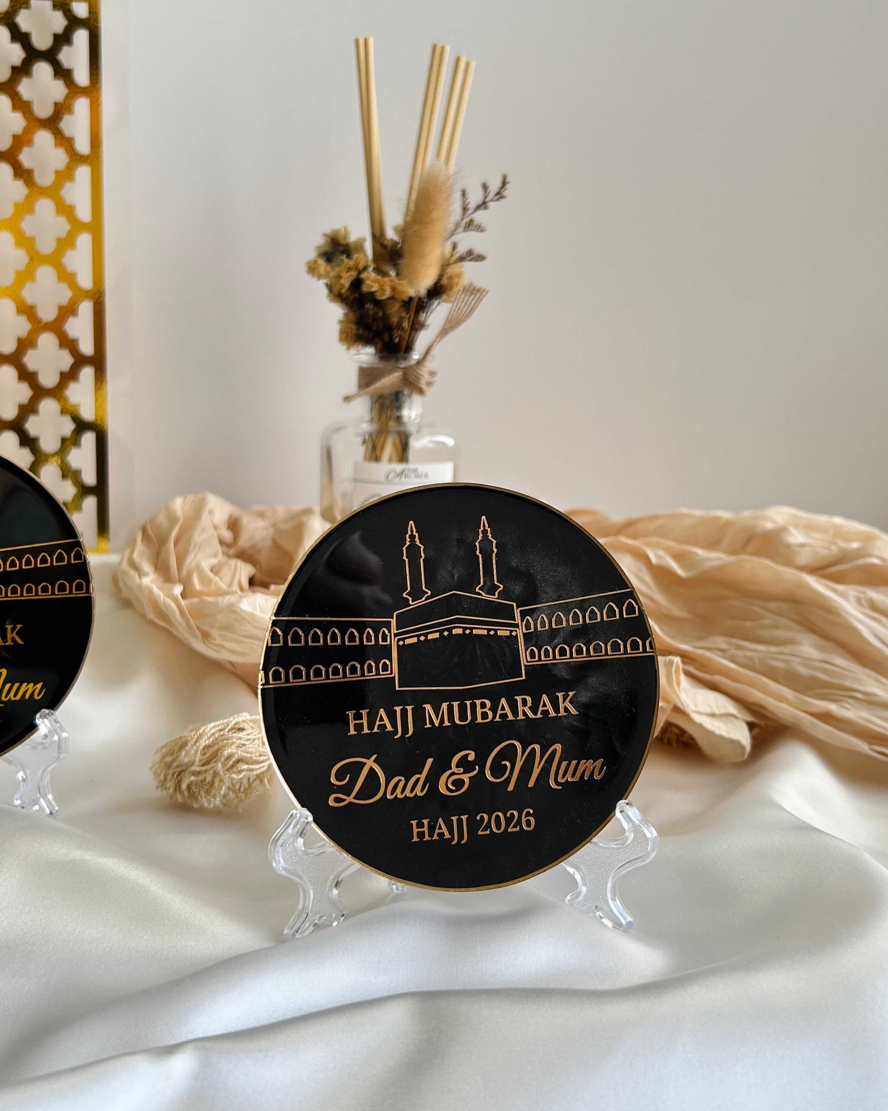 Decorative black plaque with 'Haji Mubarak Dad & Mum' text on a stand, featuring a religious symbol 'Kaaba' on a white satin background with decorative items.