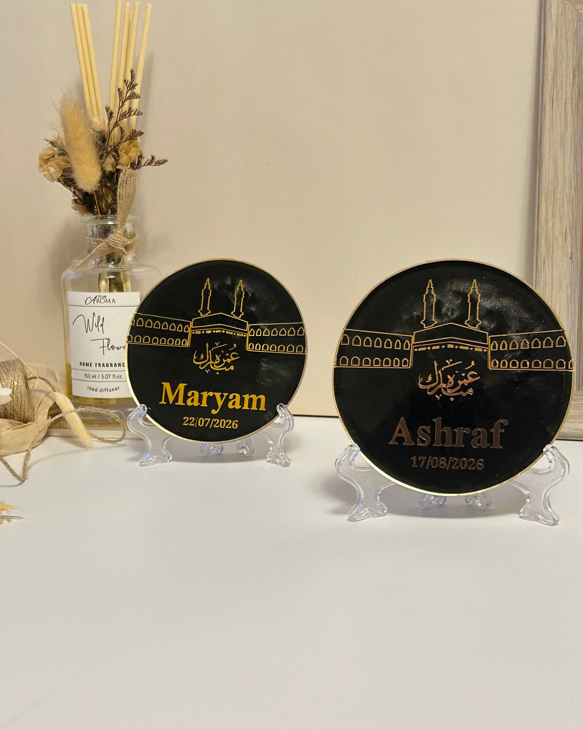 Two round black Umrah Mubarak plaque with gold Islamic line art of the Kaaba and personalised with names in gold, displayed on a clear acrylic stands on a white surface with a beige background and a decorative glass bottle behind it.