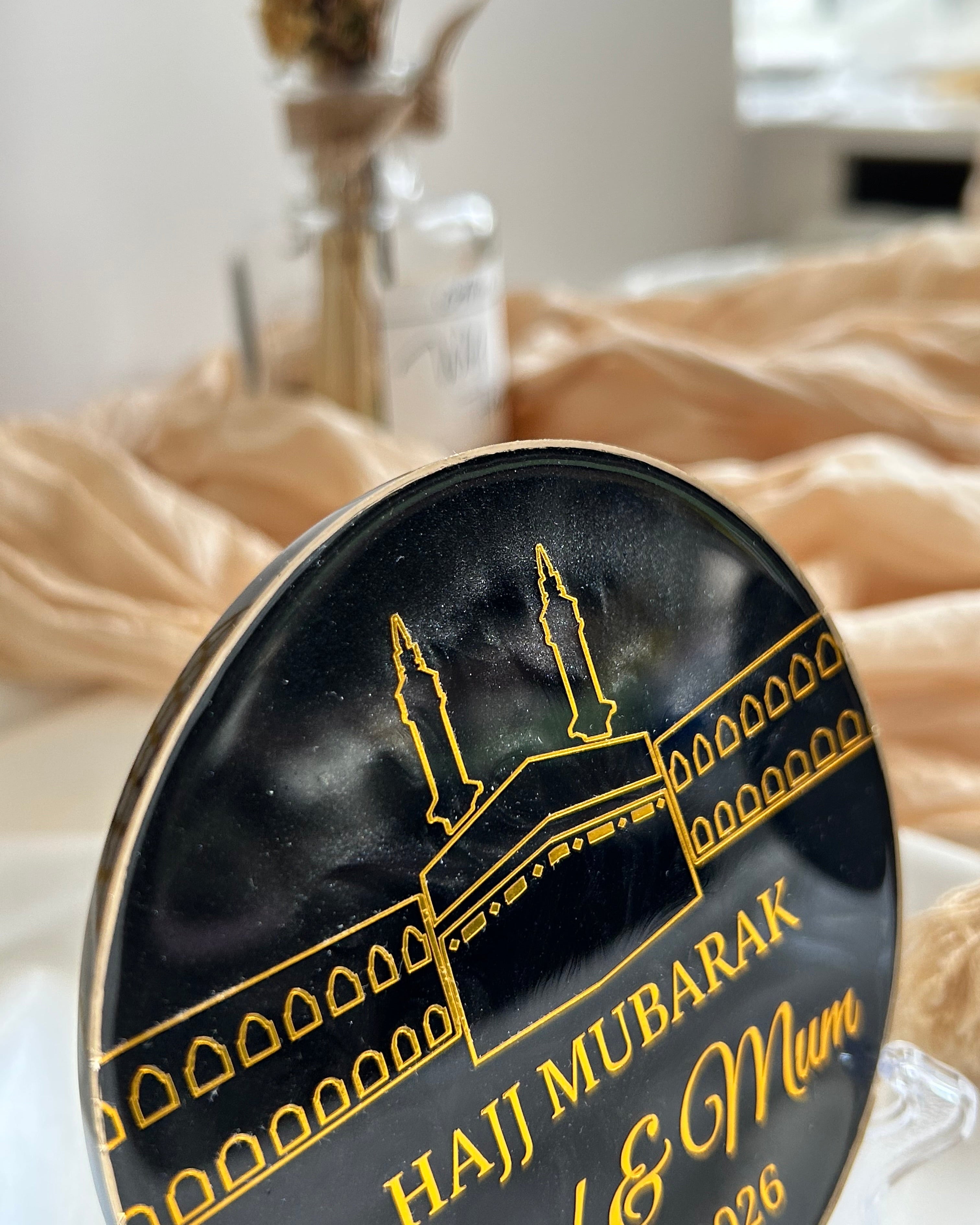 Decorative black plaque with 'Haji Mubarak Dad & Mum' text on a stand, featuring a religious symbol 'Kaaba' on a white satin background with decorative items.