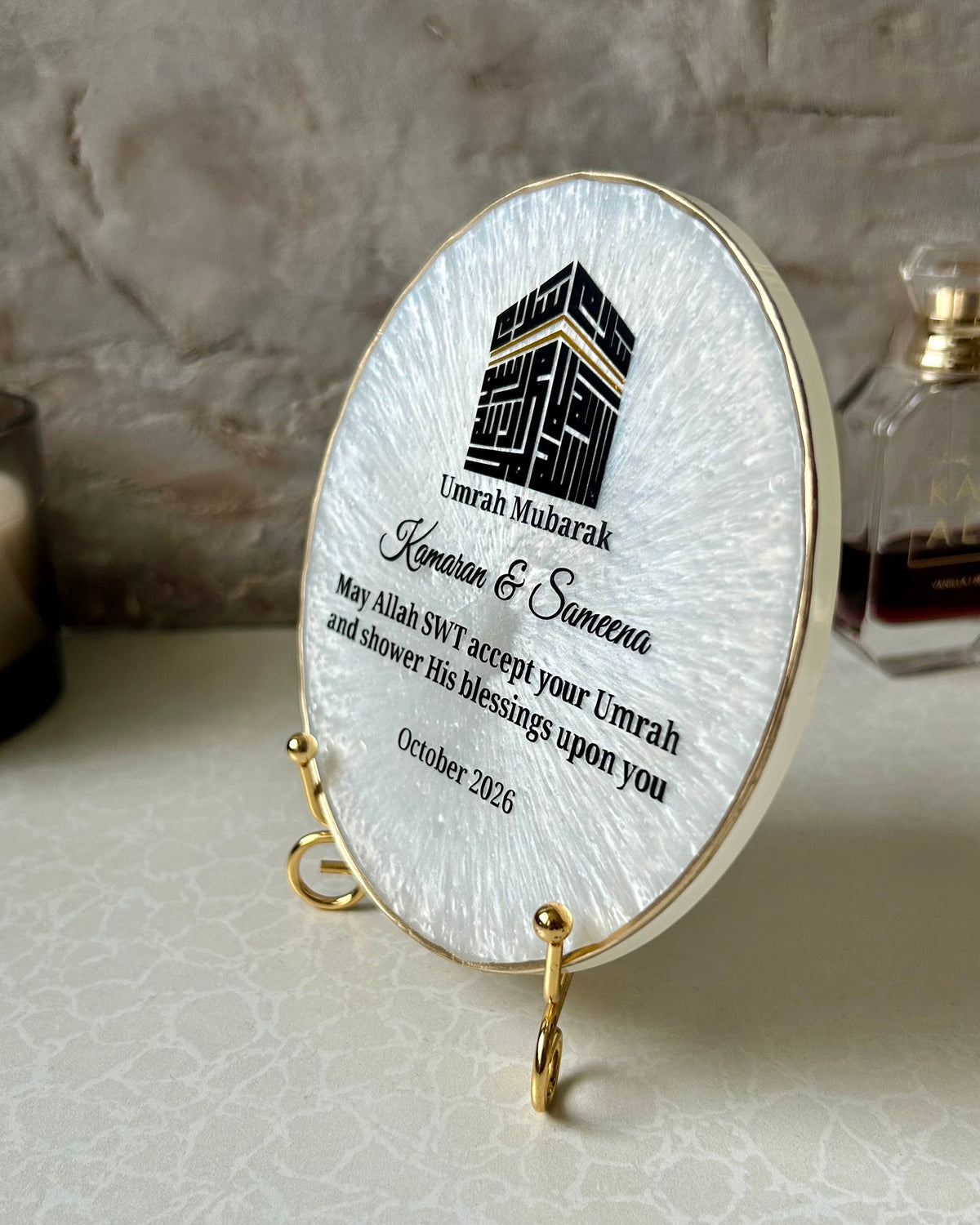 Pearlescent white Umrah Mubarak resin plaque with a gold-toned rim and display stand, featuring a black Kufic Kaaba design and text, displayed on a tabletop against a textured stone-like background.
