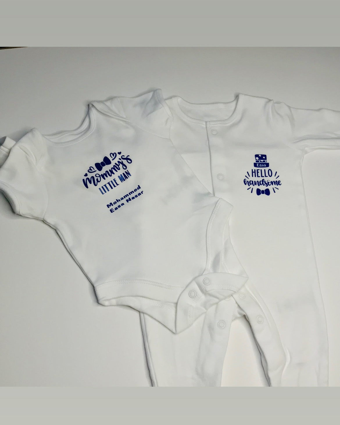 Two white baby onesies with blue text on a light gray background