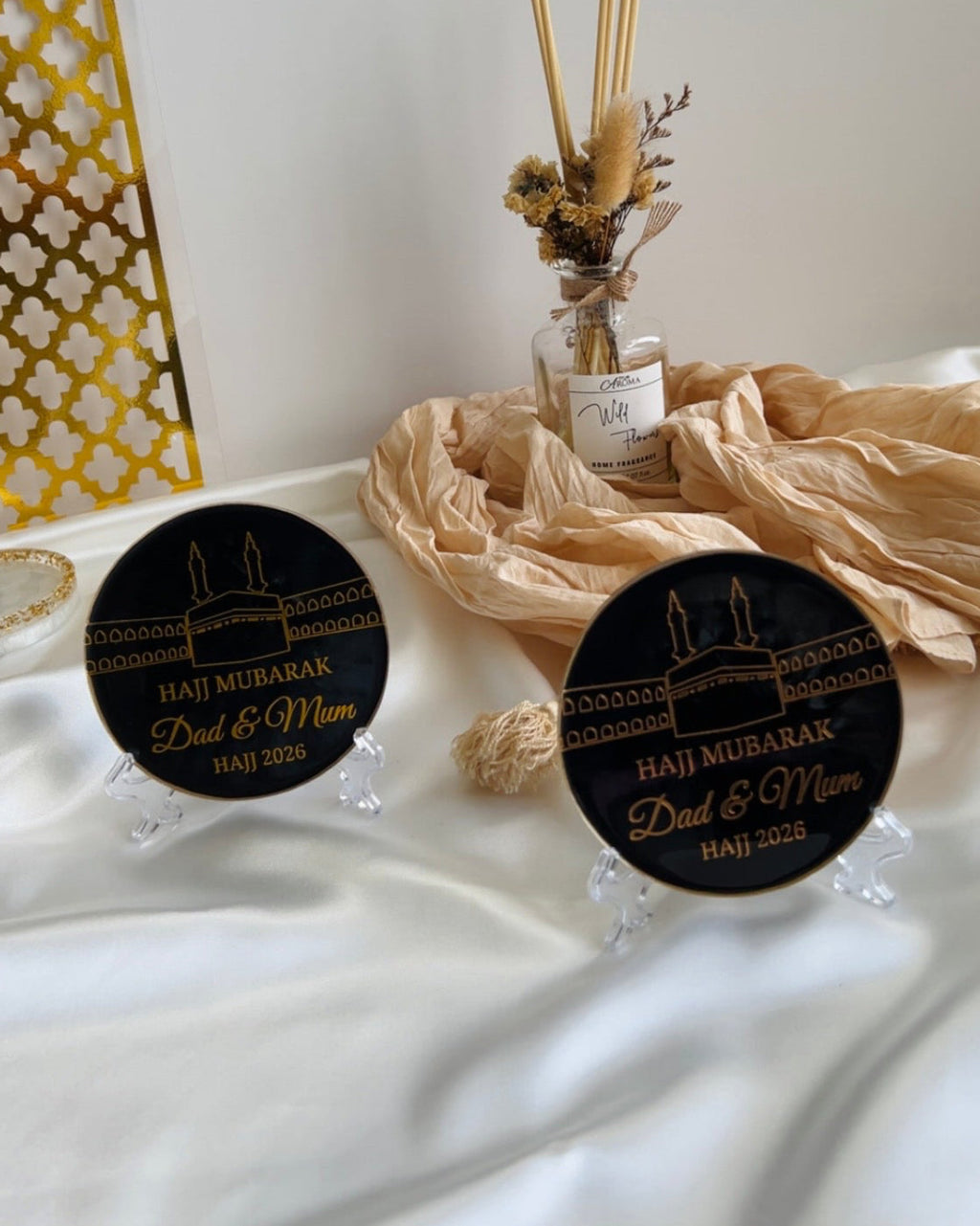 Two round decorative black plaques with 'Haji Mubarak Dad & Mum' text on a stand, featuring a religious symbol 'Kaaba' on a white satin background with decorative items.