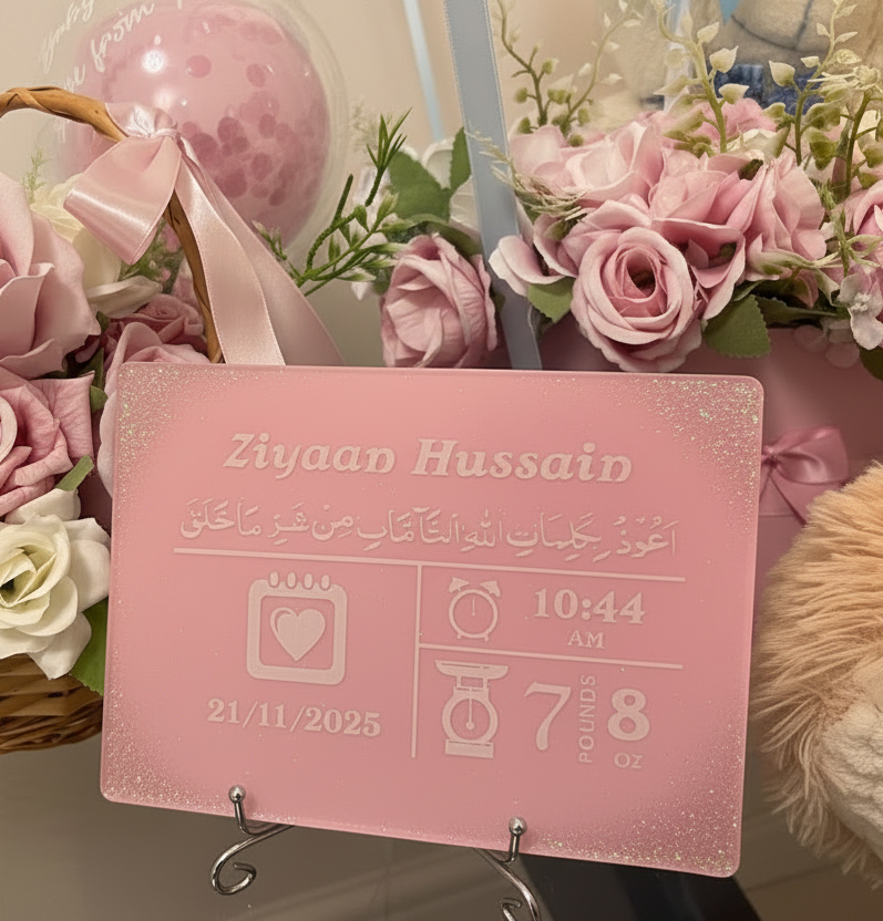 Pink baby announcement resin plaque with white personalised text resting on a silver display standing front of flowers and balloons