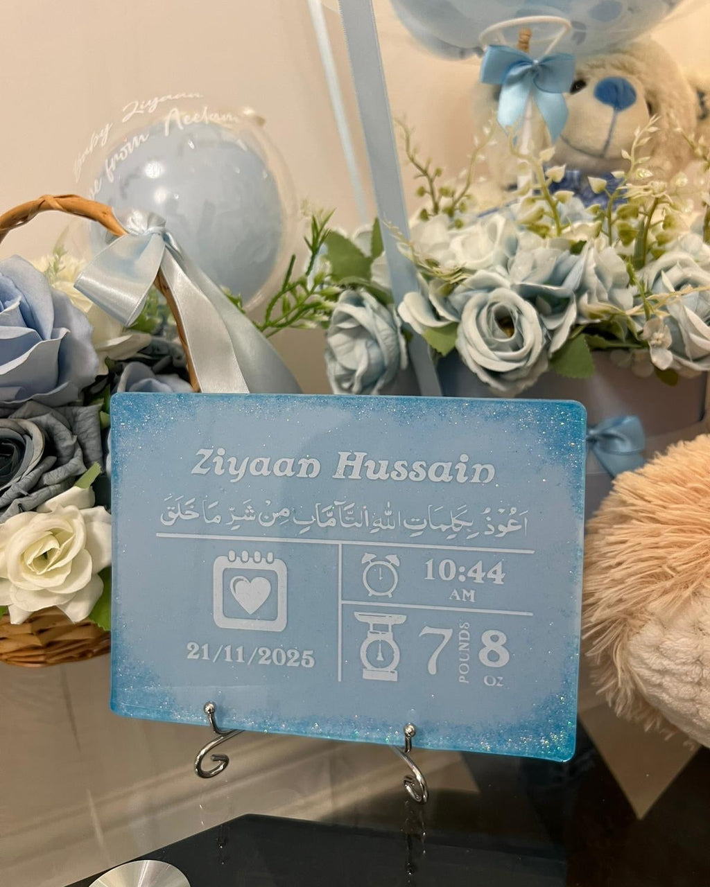 baby birth stat personalised resin plaque rectangle baby keepsake in blue with white personalised text in white and blue glitter in all for corner resting on a silver display stand on a glass table with baskets of blue flowers in the background