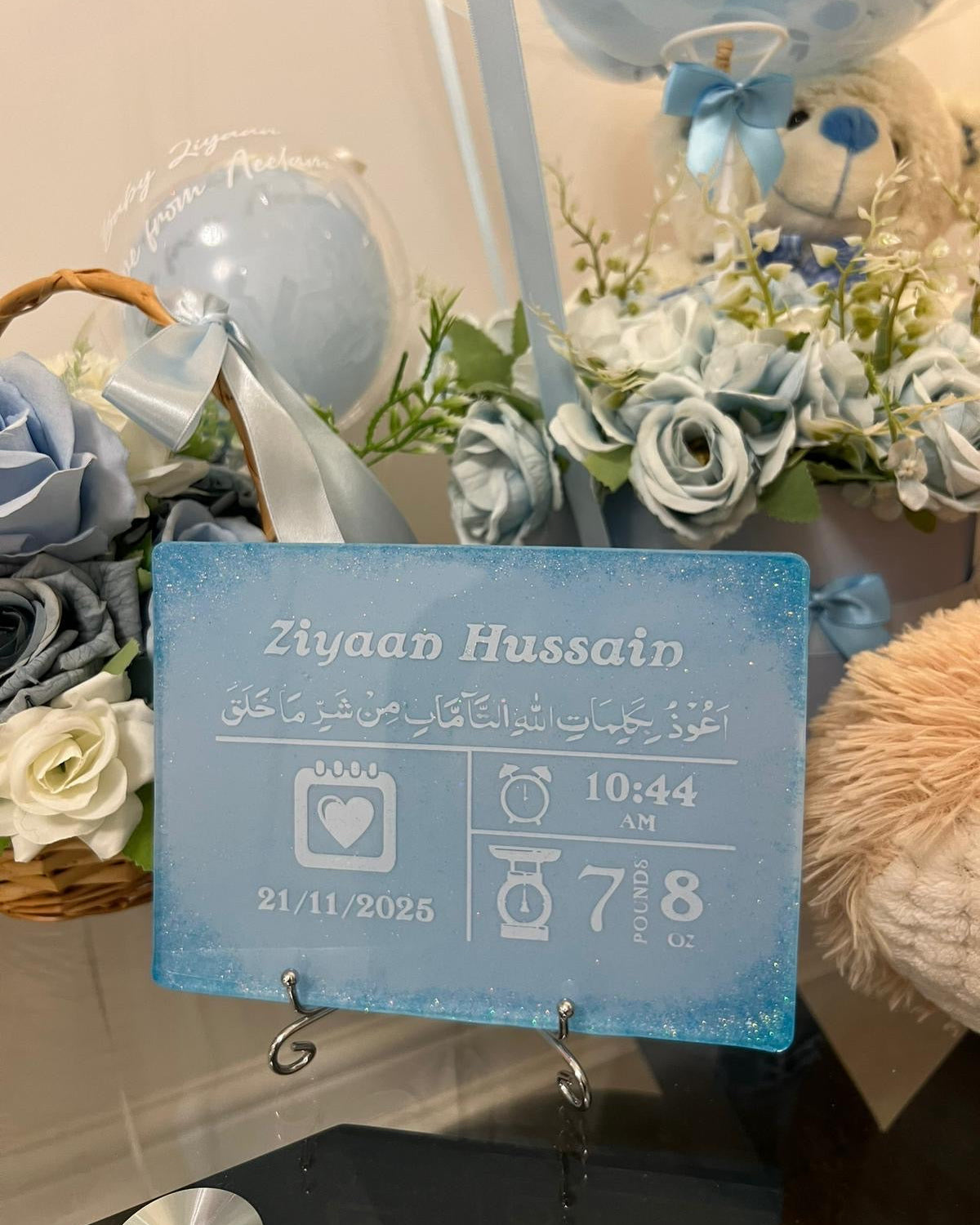 baby birth stat personalised resin plaque rectangle baby keepsake in blue with white personalised text in white and blue glitter in all for corner resting on a silver display stand on a glass table with baskets of blue flowers in the background