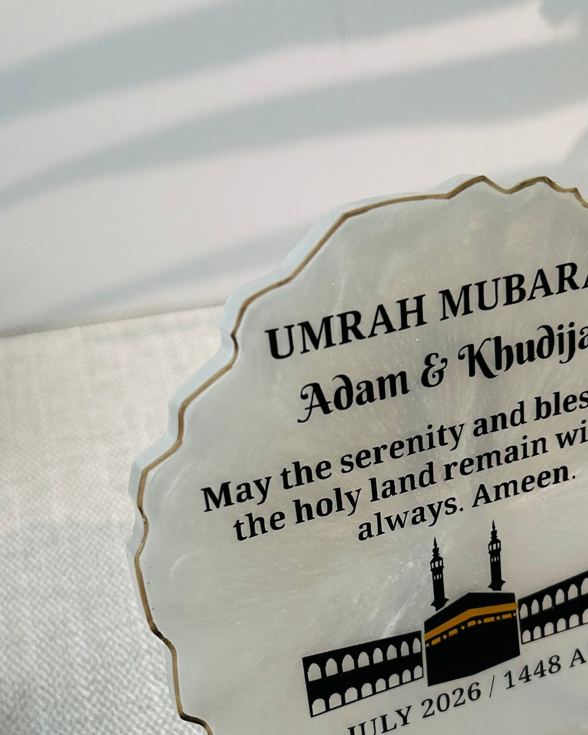 Side view of a white scalloped edge plaque with gold trim, featuring personalised text 'UMRAH MUBARAK Adam & Khadijah' and a black illustration of the Kaaba. The plaque is photographed on a light fabric surface.