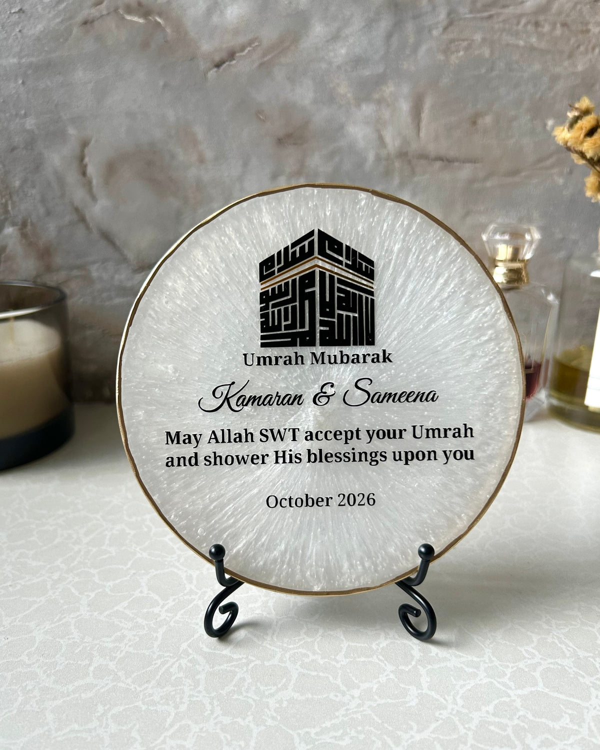 Personalised round pearlescent white Islamic plaque with gold edging displayed on a small gold stand, featuring a black Kufic Kaaba design reading Basmala and custom text for Umrah Mubarak, on a light marble surface, set against a neutral textured background.