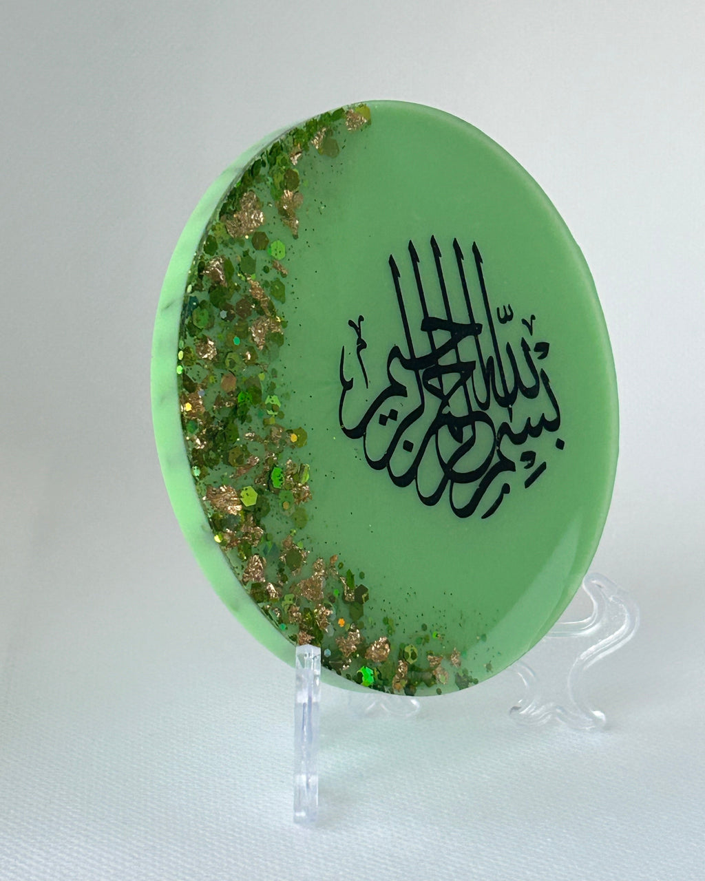 Green circular object with black Arabic calligraphy reading Bismillah Tasmiyah and gold shimmer glitter with gold leaf foiled placed sparsely displayed on a clear plastic display stand on a white background