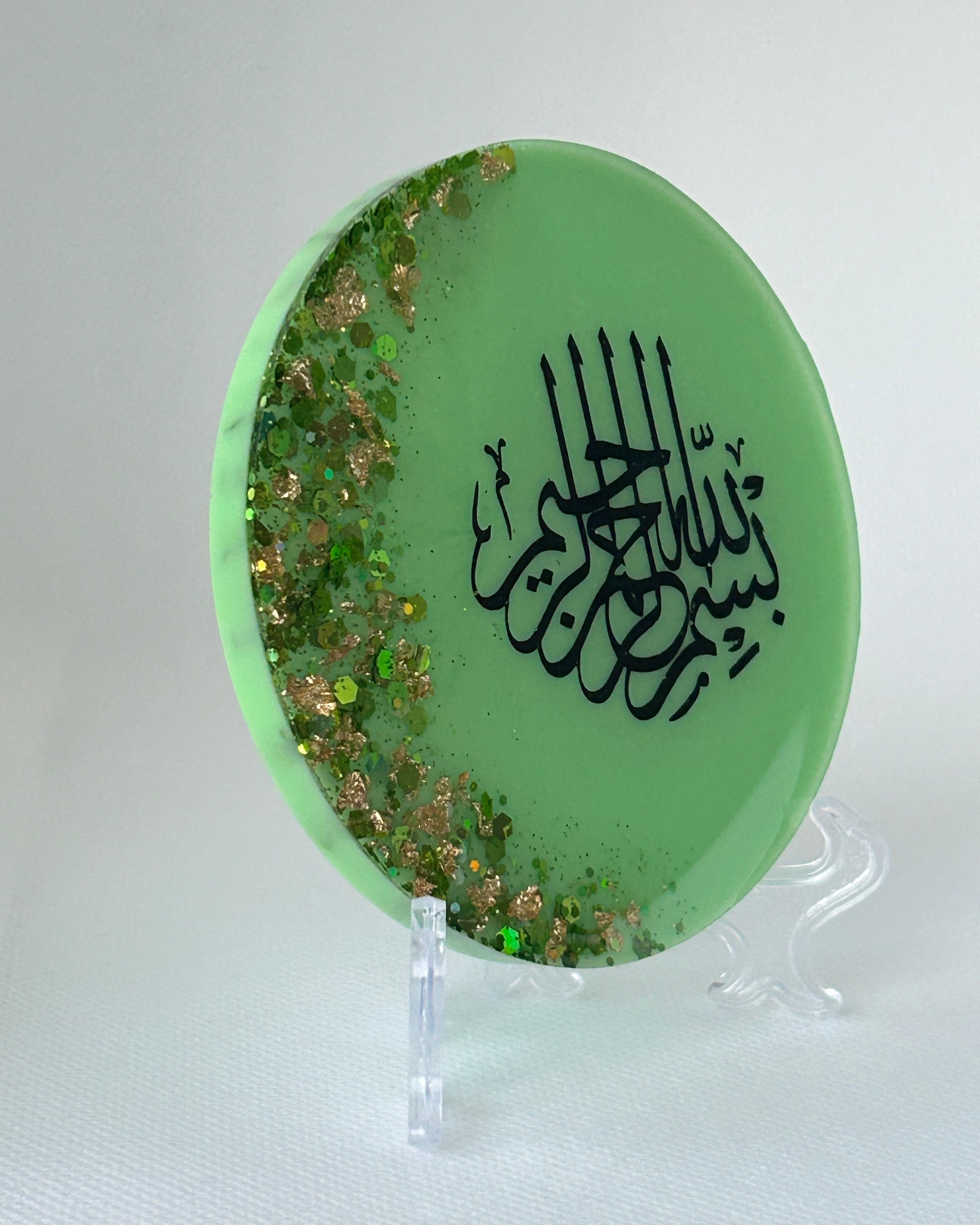 Green circular object with black Arabic calligraphy reading Bismillah Tasmiyah and gold shimmer glitter with gold leaf foiled placed sparsely displayed on a clear plastic display stand on a white background