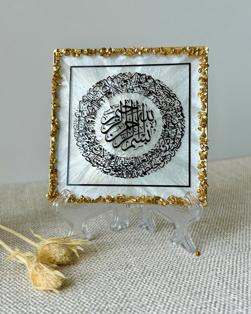 A square decorative plaque featuring black Arabic calligraphy on a white background, bordered with textured gold crushed glass trim, displayed on a clear frosted plastic display stand on a beige textured surface.