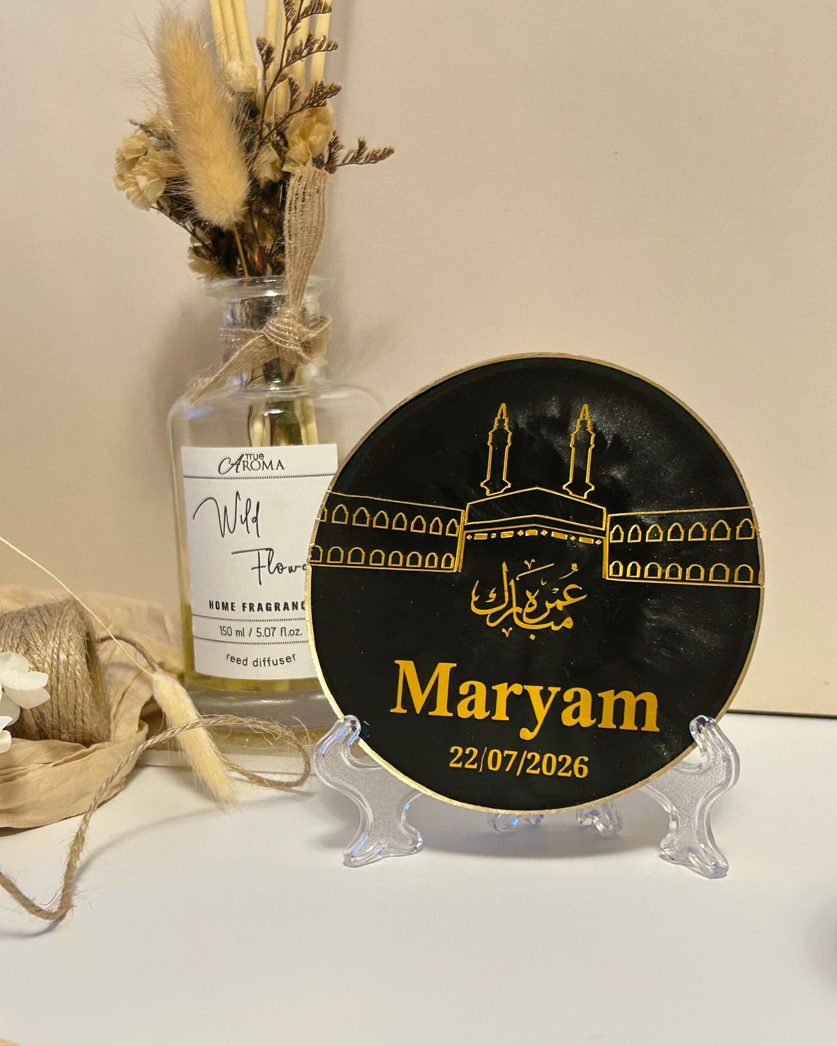 A round black Umrah Mubarak plaque with gold Islamic architecture artwork of the Kaaba and the name 'Maryam' in gold, displayed on a clear acrylic stand on a white surface with a beige background and a decorative glass bottle behind it.